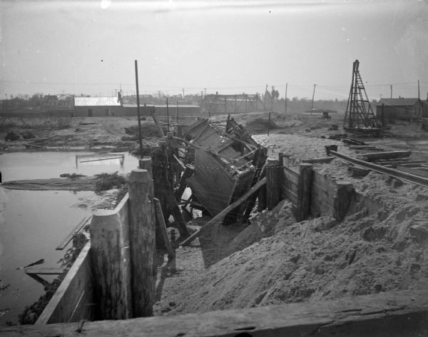 Outdoor view of a construction site, with wreckage in a ditch. Location identified as the fill and reconstruction of Town Creek after the 1911 flood in Black River Falls, and the wreck of the sand hauler.