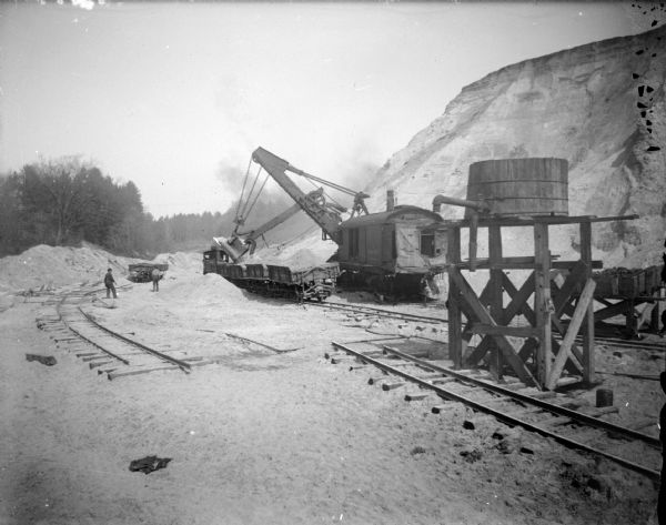 View towards a steam shovel working on a hill by a railway and water tank. Location identified as the cutting down of German Hill for fill to reconstruct the downtown of Black River Falls after the flood in October 1911.