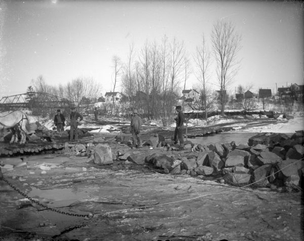 View towards men standing among several large rocks, and a road made with logs. Identified as the lying of logs on the sand bar after the flood of Black River Falls in October 1911 to aid reconstruction.