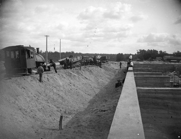 View down wall towards a construction site. On the right arec
 men by a railway track with sand haulers, and a brick viaduct. Location identified as the fill and reconstruction of Town Creek after the 1911 flood in Black River Falls.