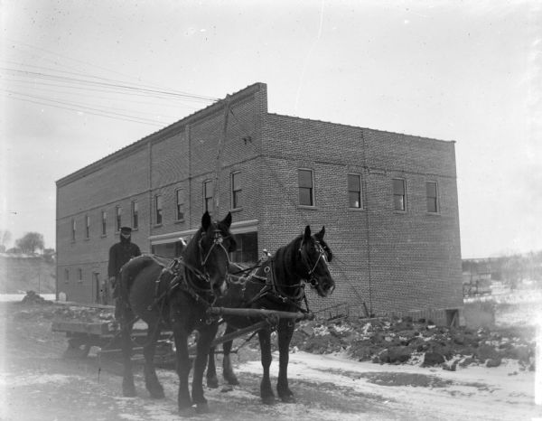 View towards a man standing on a sled behind a team of two horses on a snow-covered city street in front of a two-story brick building. Location identified as the southeast corner at the intersection of Main and First streets.