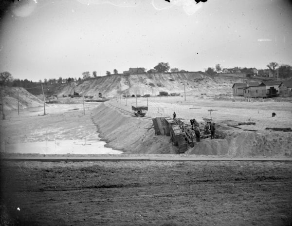 View towards a construction site, with wreckage, and men working on the wreckage. Location identified as the fill and reconstruction of Town Creek after the 1911 flood in Black River Falls, and the wreck of the sand hauler.
