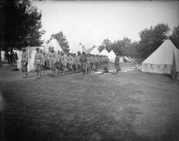 View towards a group of uniformed soldiers marching by tents. Identified as the National Guard assisting after the flood of Black River Falls in October 1911.