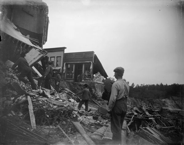 View towards several men on wreckage below several buildings. Identified as the damage after the flood in Black River Falls in October 1911, and showing the former intersection of Main and First Streets.