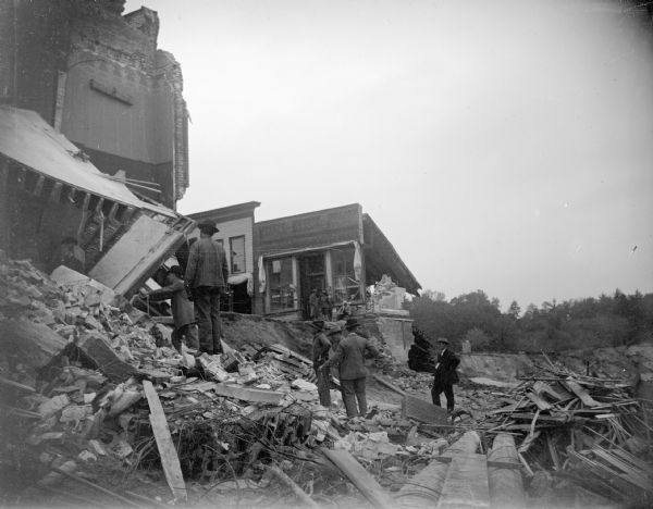 View towards several men on wreckage below several buildings. Identified as the damage after the flood in Black River Falls in October 1911 and showing the former intersection of Main and First Streets.