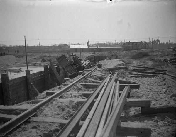 View of a railway at a construction site, with wreckage on the left side and a bridge in the background. Identified as the fill and reconstruction of Town Creek after the 1911 flood in Black River Falls, and the wreck of the sand hauler.