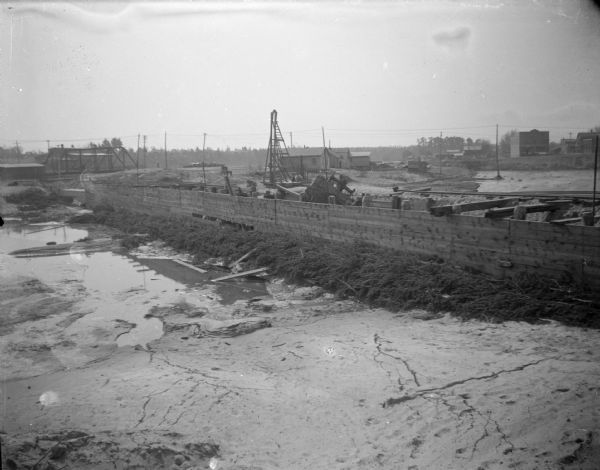 Elevated view of a construction site, with wreckage behind a wooden wall. Location identified as the fill and reconstruction of Town Creek after the 1911 flood in Black River Falls, and the wreck of the sand hauler.