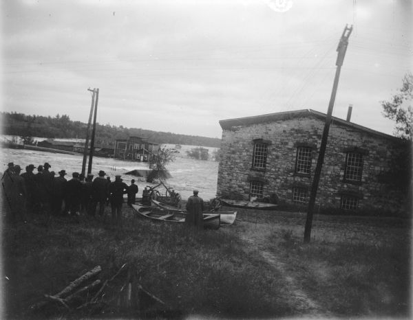 View towards river with high water flooding over several buildings behind a group of people in the foreground. Identified as the flood of the Black River in October 1911 in Black River Falls taken from Water Street on German Hill, beginning of the flood. The powerhouse is underneath the water at the center, and the Spaulding wagon shop on the right would go next in the flood. The McGillivray factory in the center would survive but would burn down in 1912.