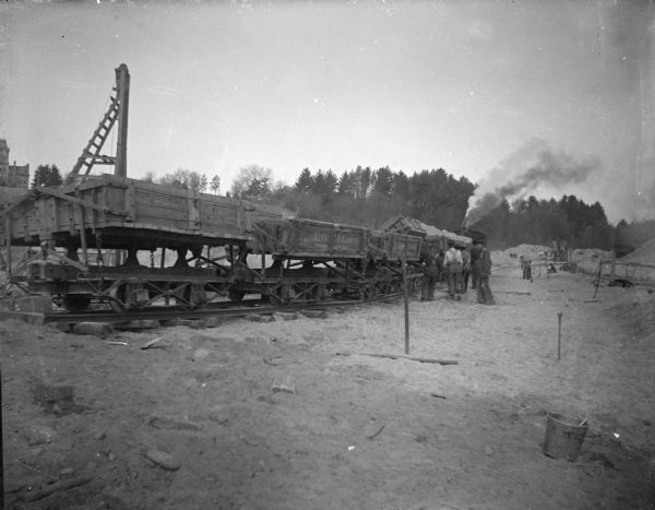 View towards men standing by a line of sand haulers on a railway. Location identified as the fill and reconstruction of Town Creek after the 1911 flood in Black River Falls.