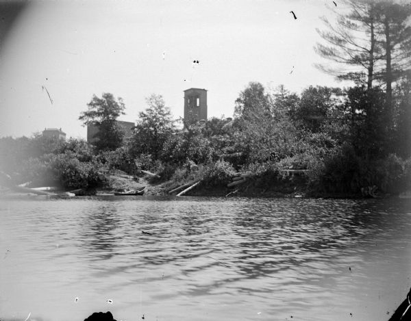 View across water towards a brick building behind a group of trees along the shoreline. Identified as the York iron mine and boarding house.