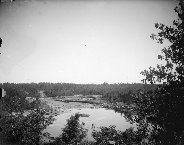 Elevated view over trees towards large groups of logs in a river. Identified as the Black River.