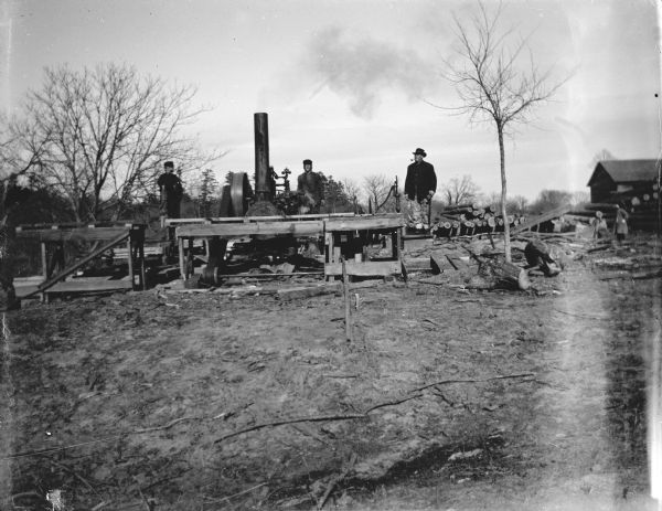 View towards three men posing standing near a steam-powered open saw mill next to piles of logs.