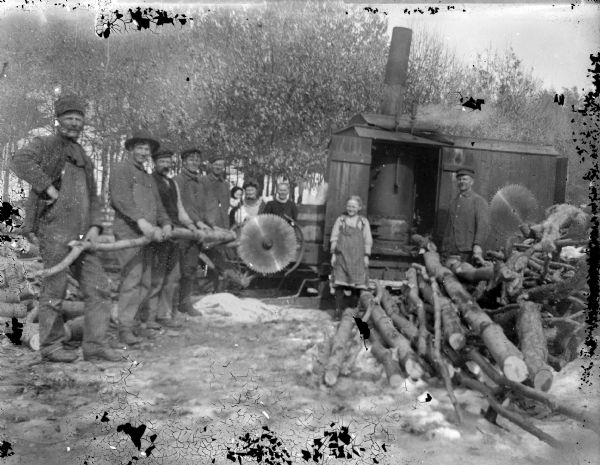 Group portrait of men, women, and a child posing standing next to a steam-powered open saw mill next to piles of logs.