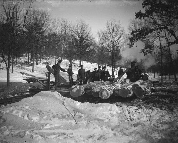 View towards a group of men posing standing by a pile of logs on snow-covered ground in front of a steam-powered saw mill.