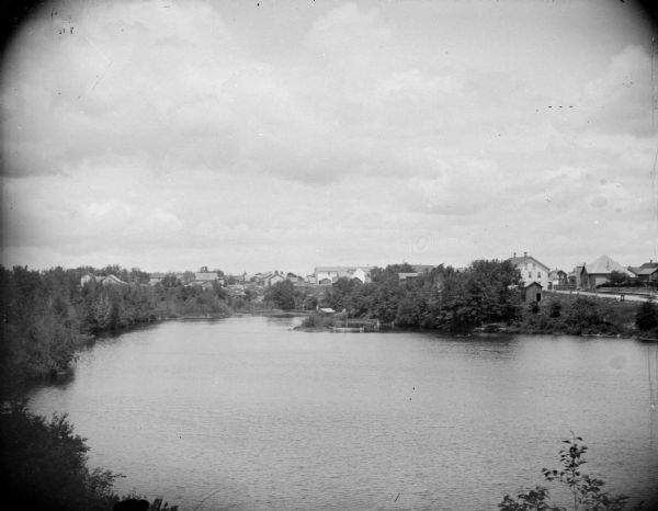 Elevated view across water towards a town on the far shoreline. Identified as the town of Merrillan taken from the bandstand of Oakwood Park looking north over Oakwood Lake. The chapel is on the right, and on the left across Main Street is the Merrillan House. The long building located in the center is the Trow Store.