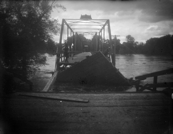 View from shoreline towards a damaged bridge across a body of water. Identified as the bridge across the Black River at Melrose. A man is standing at the end of the bridge near a broken section.