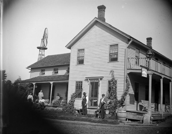 View towards men and women posing sitting and standing in front of a wooden building. The top of a windmill behind the building rises above the roof. Identified as the North Bend Hotel in Melrose.
