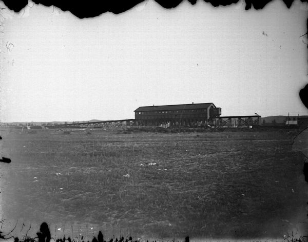 View across field towards a long building in the distance by an open field. Identified as the railroad coal shed in Millston.