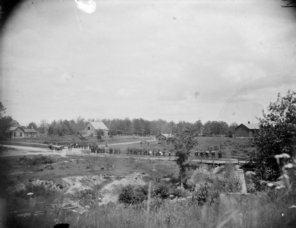 View across field towards a large group of people in the distance walking behind a band along on a town street. Identified as the reunion of the Grand Army of the Republic in June 1895 in Merrillan, marching south on Hammond Street near the corner of Mill Street to a camp meeting at Oakwood Park. Hall's Creek is in the foreground.