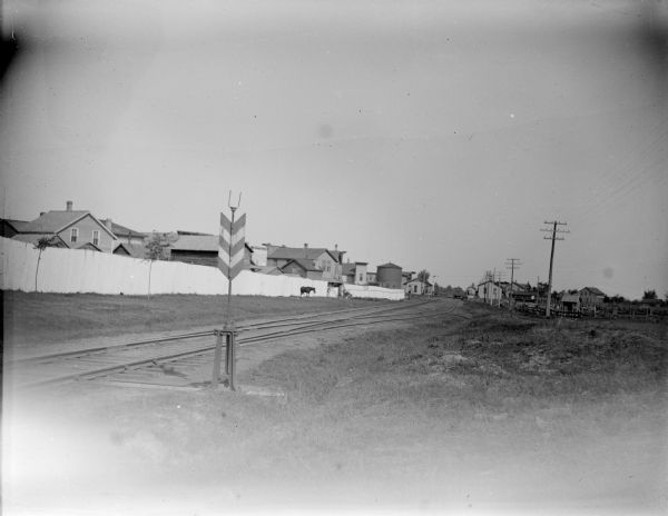 View across railway tracks near a town. Identified as the town of Merrillan, looking north from the Pearl Street crossing. The buildings on the other side of the long fence are probably the rear of buildings of East Main Street between Pearl and Omaha Streets. The water tank in the distance is at the depot.