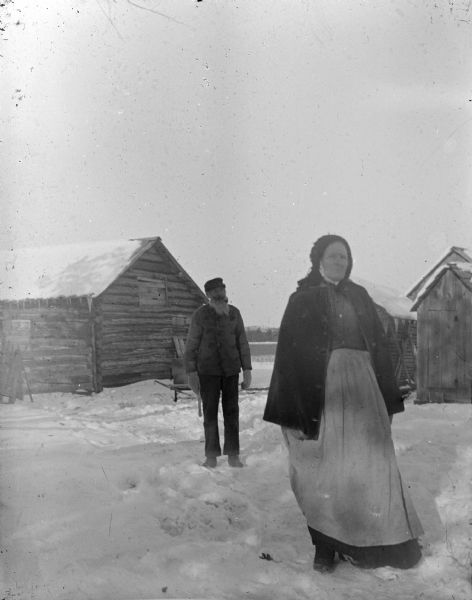Outdoor portrait of an elderly woman and man posing standing on snow-covered ground in front of wooden buildings.