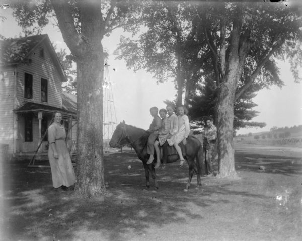 Outdoor group portrait of four children posing sitting on a horse. A man and woman are posing standing on either side of the horse. In the background are trees in the yard of a wooden house.