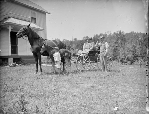 Outdoor group portrait of two women posing sitting in a buggy pulled by a single horse. A boy is standing and displaying a single foal. On the right a man is standing near the buggy. The group is in the yard of wooden house. A dog is rolling in the grass near the porch.