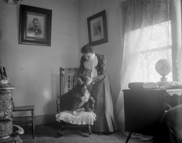 Portrait of a woman posing standing and petting a dog that is sitting on a chair near a window. A woodburning stove is in the foreground on the left. There are portraits on the walls.