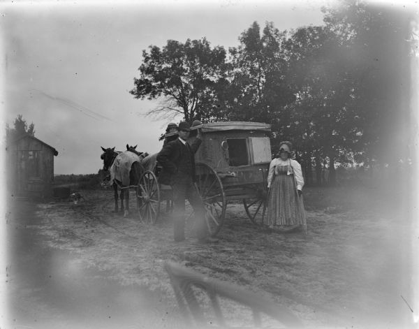 Outdoor portrait of a man and woman posing standing by the rear of a wagon pulled by a team of two horses. There is a man posing sitting in the wagon. Identified as a peddler's wagon.