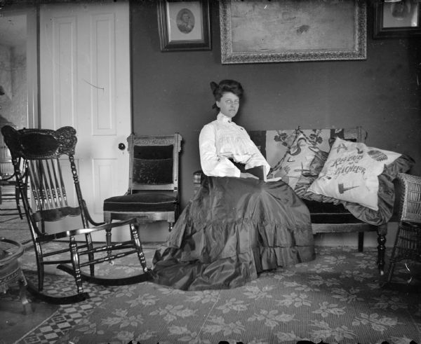 Indoor portrait of a woman posing sitting on a sofa. She is wearing a dark-colored skirt and light-colored blouse, near a pillow that reads: "Reveries of a Bachelor."