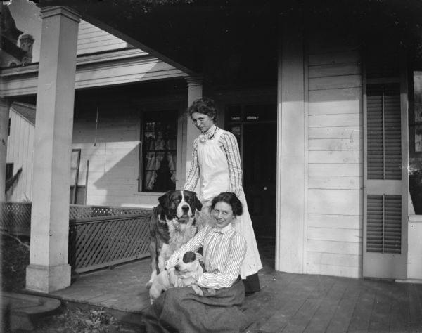 Portrait of a woman posing sitting on a porch holding a small dog, and another woman posing sitting next to a large dog. Both women are wearing eyeglasses.