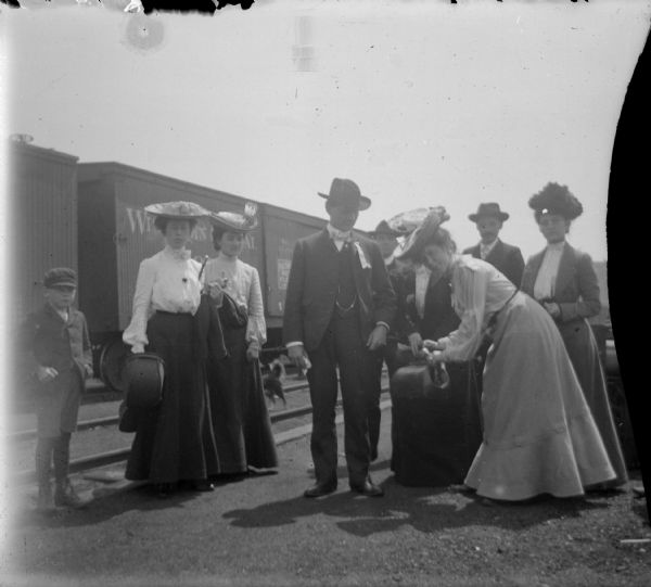 Portrait of a group of men, women, and a child on a railroad platform. Railroad cars are in the background.