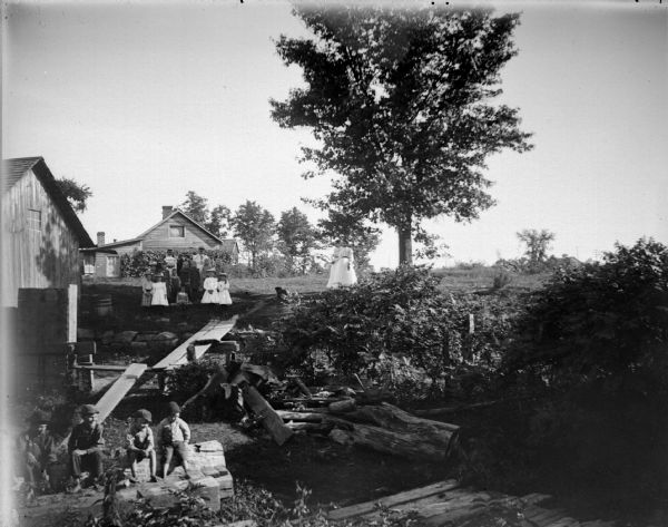 Slightly elevated view of four boys posing sitting on a log in the foreground, and on the hill behind them, a large group of women posing together sitting and standing in the background. Two women are posing standing on the right. The group of people is in a yard with several wooden buildings.