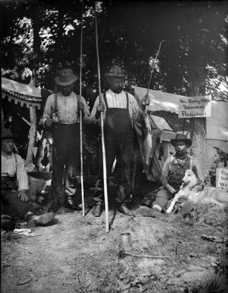 Group portrait of two men posing standing and holding fish and fishing poles, and two men are sitting on the ground on either side of the standing men. The man on the right is sitting with a dog in his lap. A sign on the tree on the right reads: "The Weary Willies Summer Residence."