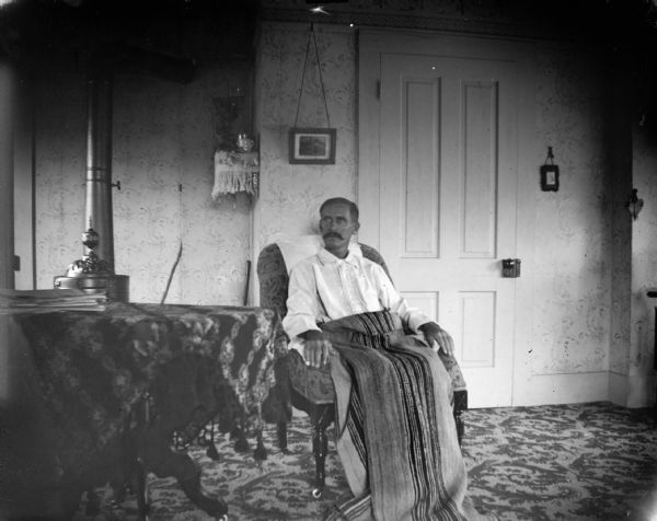 Interior portrait of a man sitting in a chair with a blanket over his lap, next to a draped table. There is a closed door behind him.