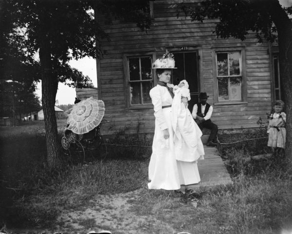 View towards a woman wearing a light-colored dress, posing standing and holding an infant in her arms while standing in the yard of a wooden house. There is a man sitting on the steps in the background. A girl is standing next to a tree on the right.