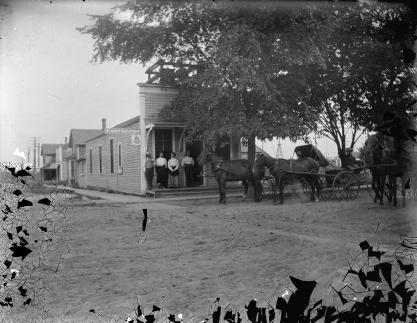 View across unpaved street towards two men and two women posing standing on the porch of a wooden storefront building. Three wagons and a horse are on the right. Location identified as Warrens in Monroe County.