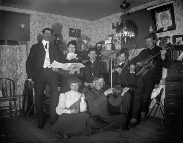 Interior portrait of five men and two women posing sitting and standing in a room holding musical instruments and sheet music.