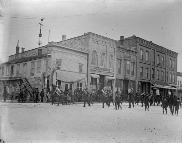 View across street towards a marching band coming up a snow-covered street. People are watching from the wooden sidewalks, and young boys are walking along with the band. Identified as the intersection of Main and First streets, at the northeast corner. A sign above one of the storefronts reads: "A. Meinhold."