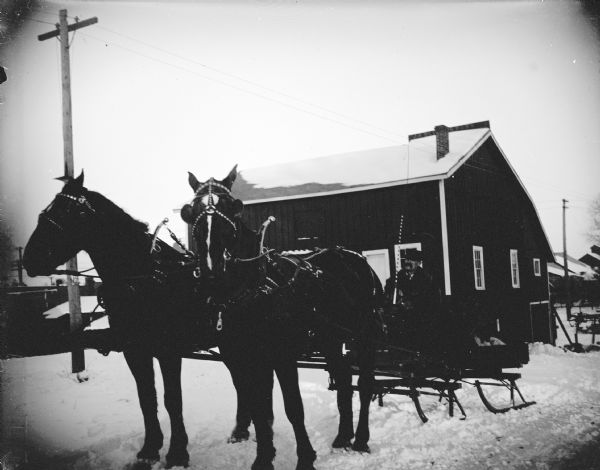View towards a man sitting in a sleigh pulled by a team of two horses on snow-covered ground in front of a wooden building. The building is identified as the Price barn, owned for a stagecoach line on the northeast corner of Harrison and Third streets. Later Abe Bailey owned the building.