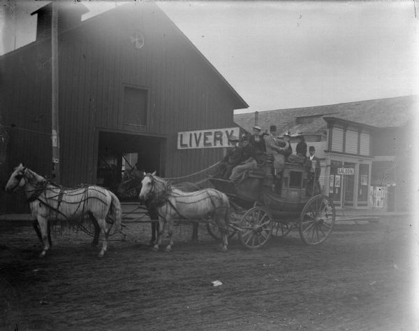 View across unpaved street towards a group of people posing sitting on a stagecoach pulled by a team of four horses wearing flynets. The stagecoach is in front of a livery stable and saloon.