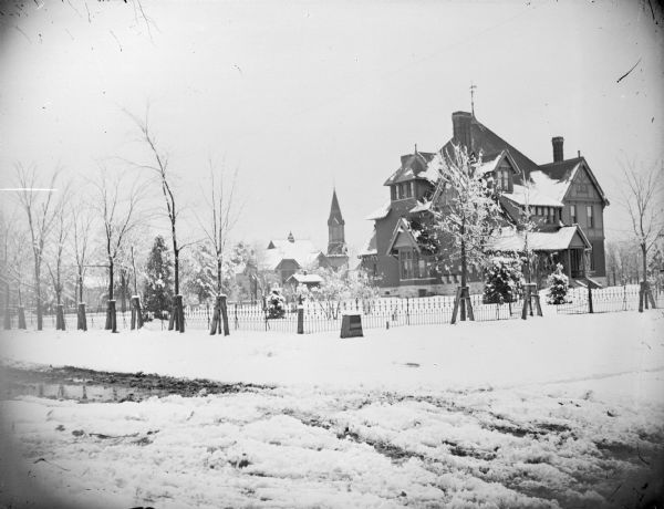 View from snowy street towards a large house surrounded by a snow-covered yard. Identified as the house of the Price family built in 1881, which occupied an entire city block in Black River Falls between Fourth and Fifth streets, and Harrison and Tyler streets.