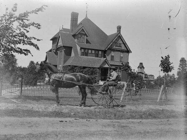 View across road towards a man sitting in a buggy in front of the fence surrounding a large house. Identified as the house of the Price family built in 1881. The house occupied an entire city block in Black River Falls between Fourth and Fifth streets, and Harrison and Tyler streets.