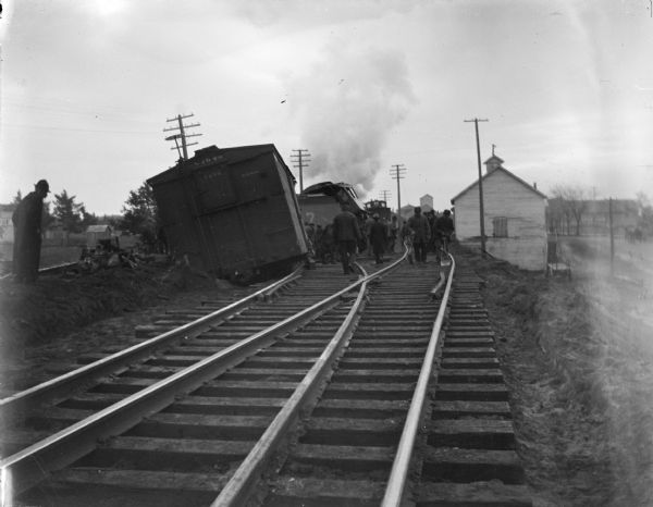 View down railroad tracks towards a large group of people standing near the wreck of a train. Railroad cars have fallen over to the left of the tracks. Two men are working with shovels on one set of the tracks on the right. There are buildings in the background.