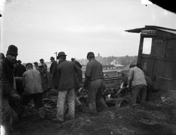 Close-up of a group of men cleaning up the wreck of a train. Pieces of railroad cars are in piles of dirt. A railroad car is on the right. A town is in the background.