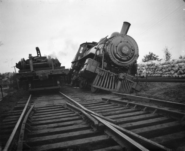 View down railroad tracks towards the front of a locomotive tipped sideways on the set of railroad tracks on the right. On the left set of railroad tracks is a railroad car carrying parts on its open bed. In the background on the right is a pile of sacks.