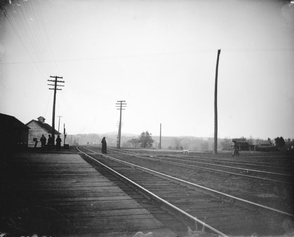 View down railroad platform towards men waiting near a power line on the left. Railroad tracks stretch from the platform and curve off to the right near a town in the distance. A person is walking on a path across the railroad tracks in the center, and a dog and a woman are approaching the tracks from the right.