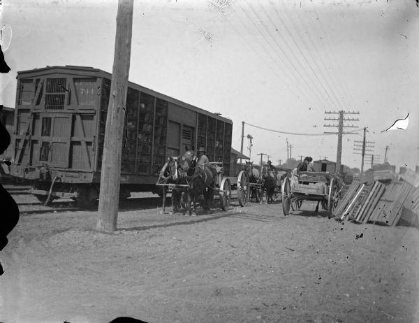 View towards three horse-drawn wagons to the right of a railroad boxcar.