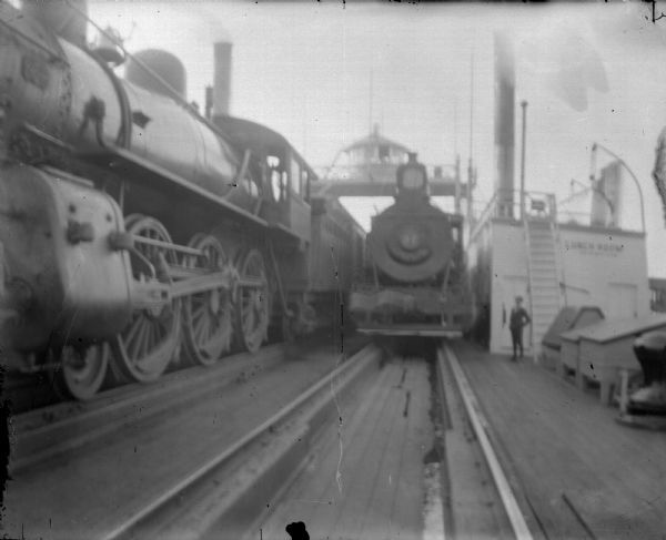 View down center of railroad tracks towards a train yard, showing several trains with locomotives. A man is standing near a building on the right has a sign that reads: "Lunch Room." Image not in focus. Identified as probably not local.