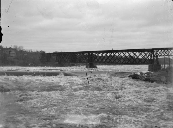 View looking upriver towards an individual in the distance crossing a bridge across a river. Location identified as the bridge across the Black River at Black River Falls. Buildings are on the far shoreline on the left.